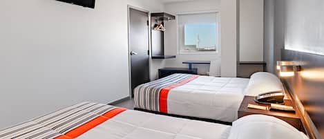 A hotel room with two beds, a flat-screen TV, a small desk with a lamp, and a window with a view of a tower.