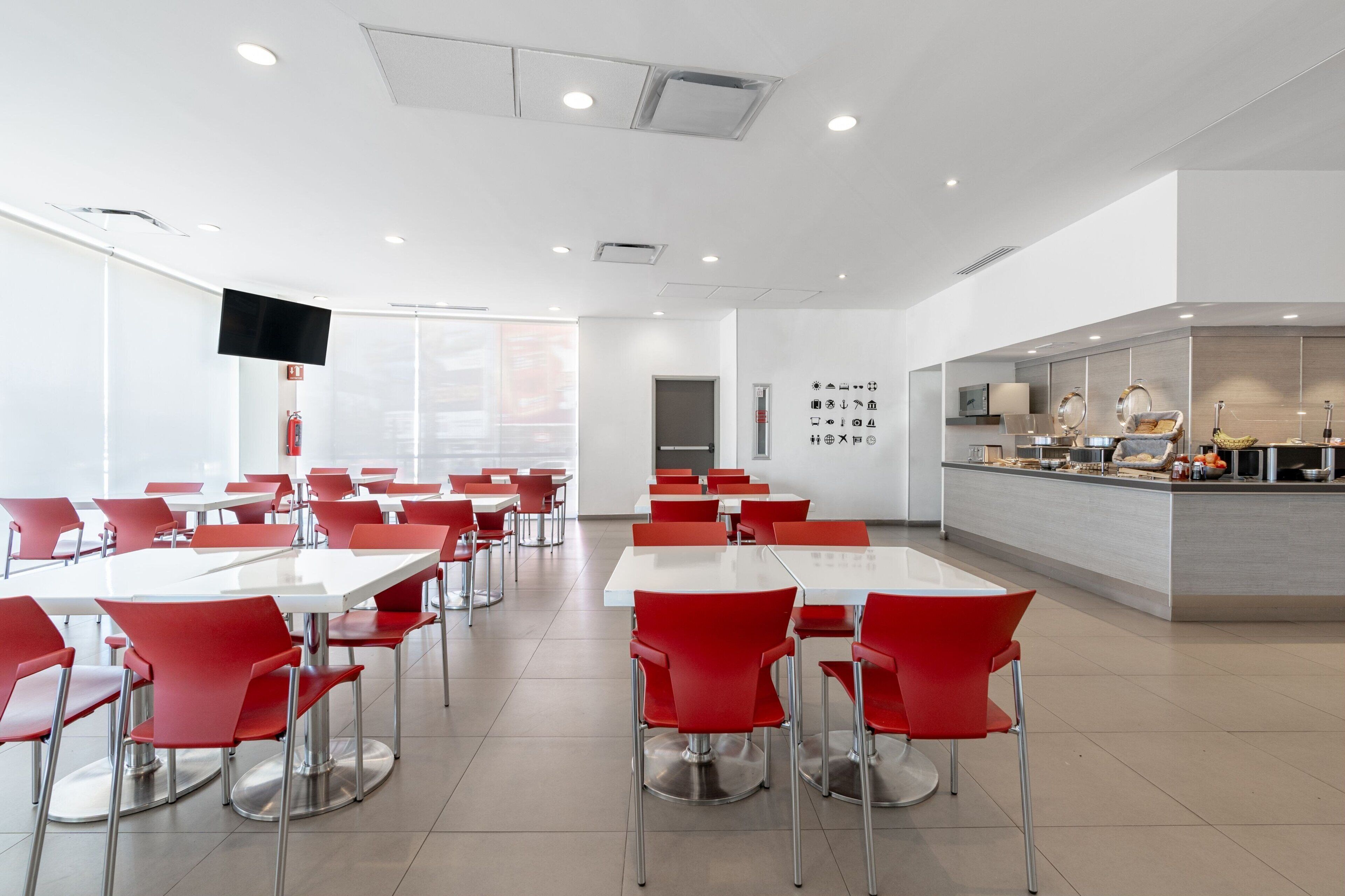 A modern dining area with red chairs and white tables, a kitchen counter with food items, and a flat-screen TV mounted on the wall.