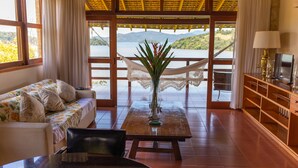 A room with a sofa, a table with a vase, a TV, and a view of a lake and hills. - Santa Clara Hotel (Paraty)