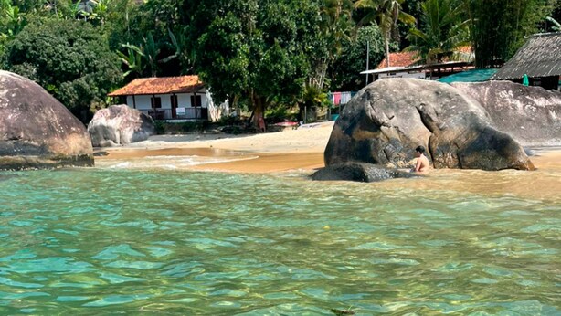A beach with clear water, large rocks, and a hut in the background. - Santa Clara Hotel (Paraty)