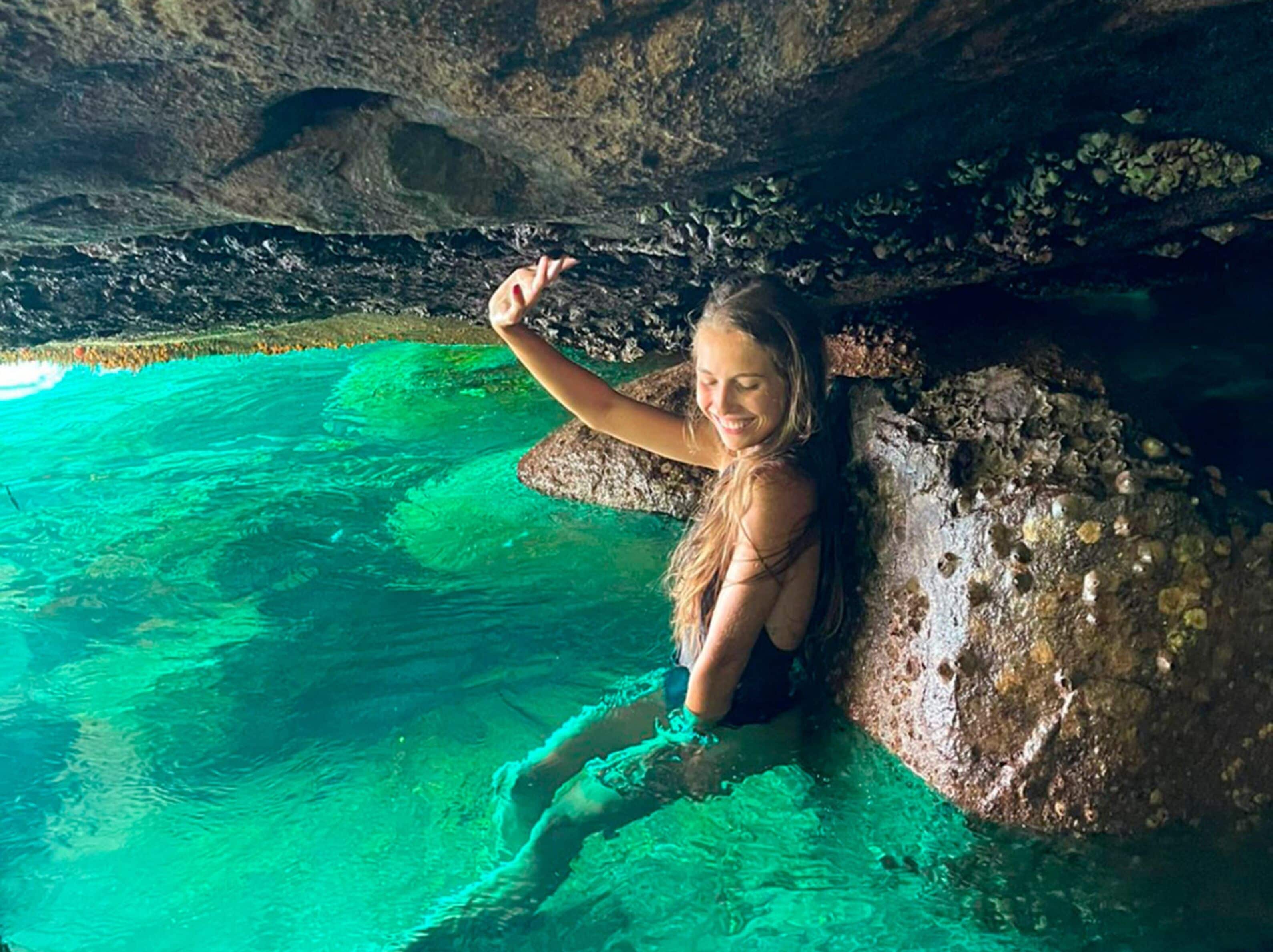 Una persona sentada sobre una roca junto a una piscina azul cristalina con un techo que recuerda a una cueva.