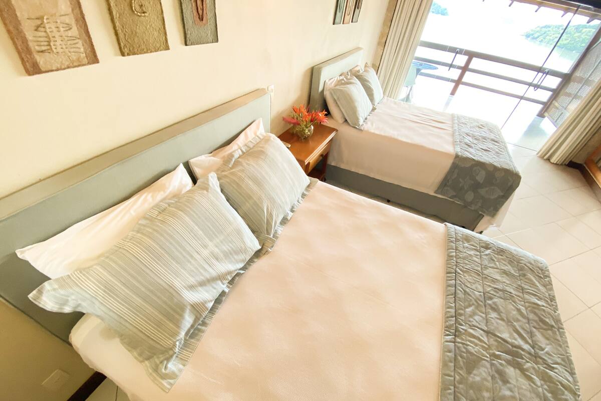 A hotel room with two beds, a wooden headboard, a small table with a vase of flowers, and a view of the outdoors through large windows.