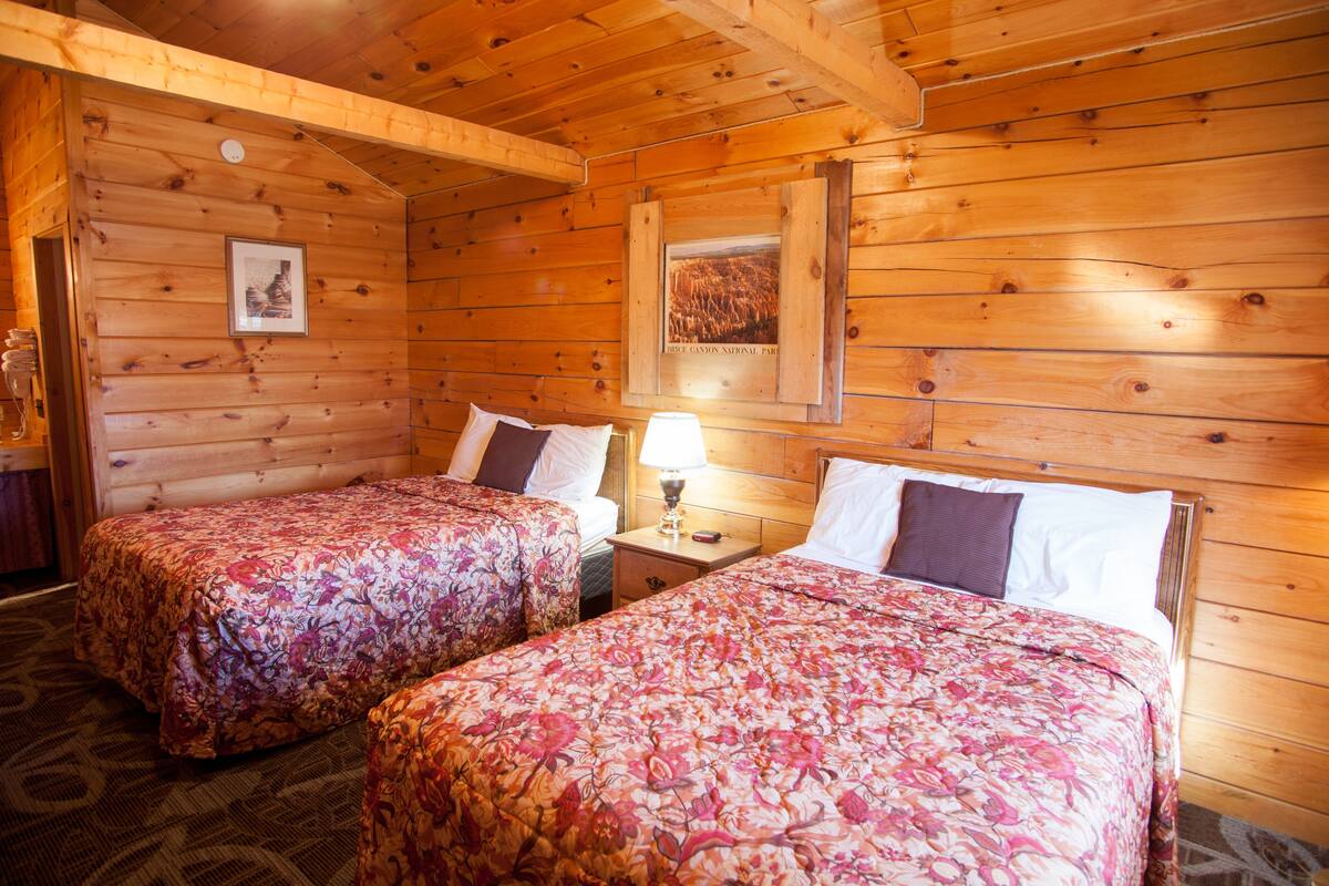 Quarto de cabana de madeira com duas camas, uma mesa de cabeceira, um abajur e quadros nas paredes.
