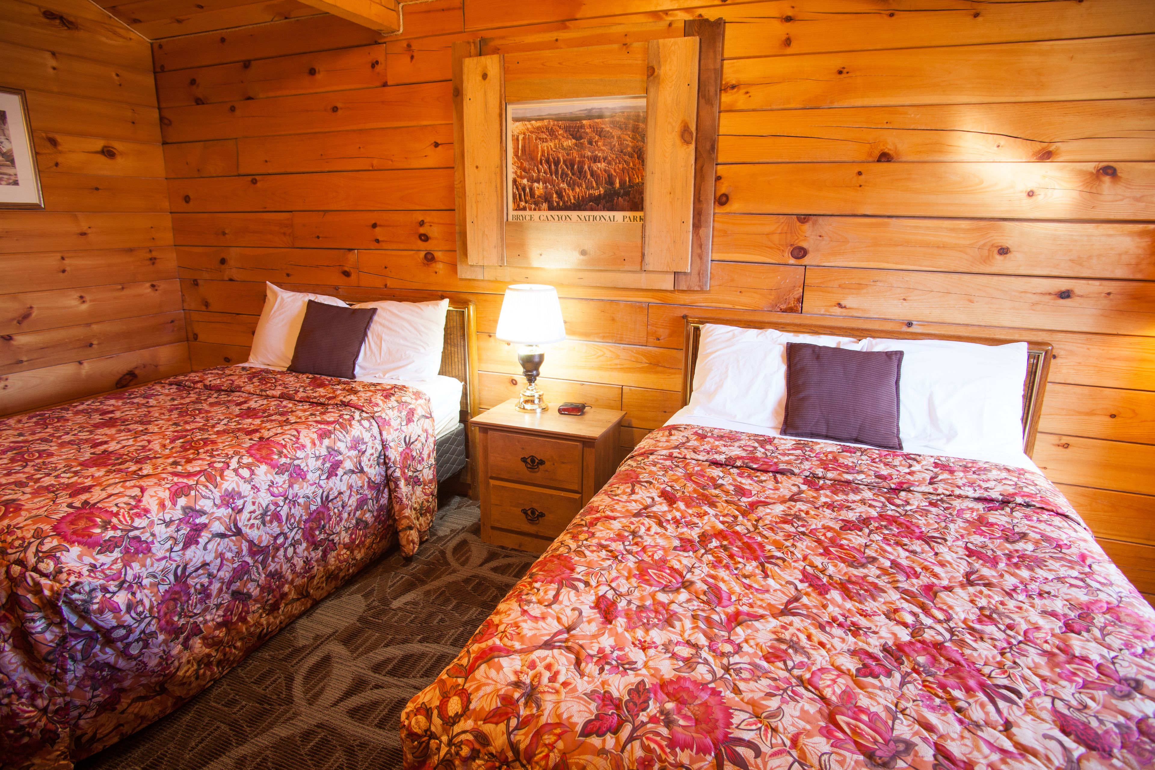 A wooden cabin room with two beds, a nightstand with a lamp, and a framed picture on the wall.