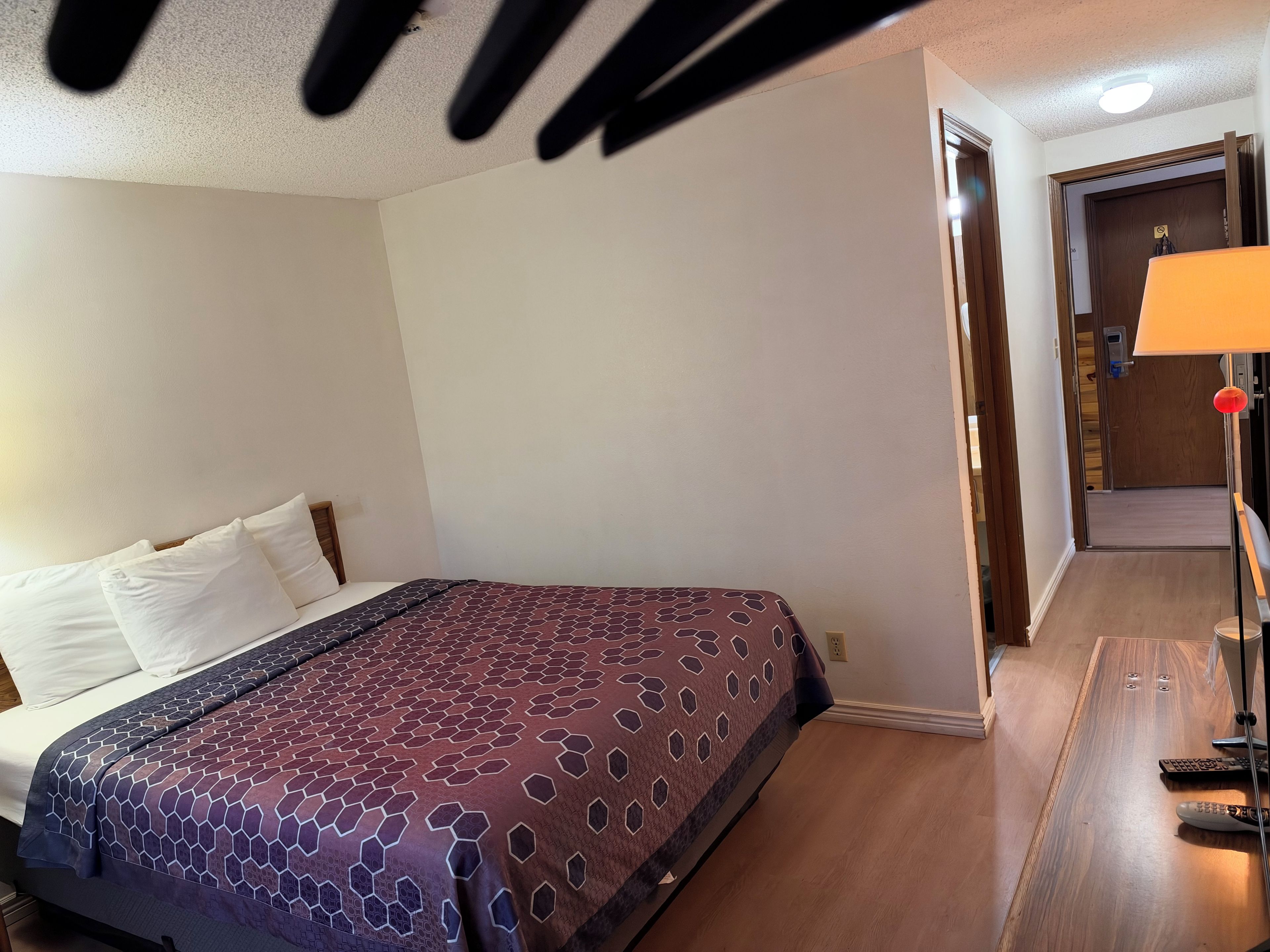 A hotel room with a bed, a wooden floor, a door, and a lamp.