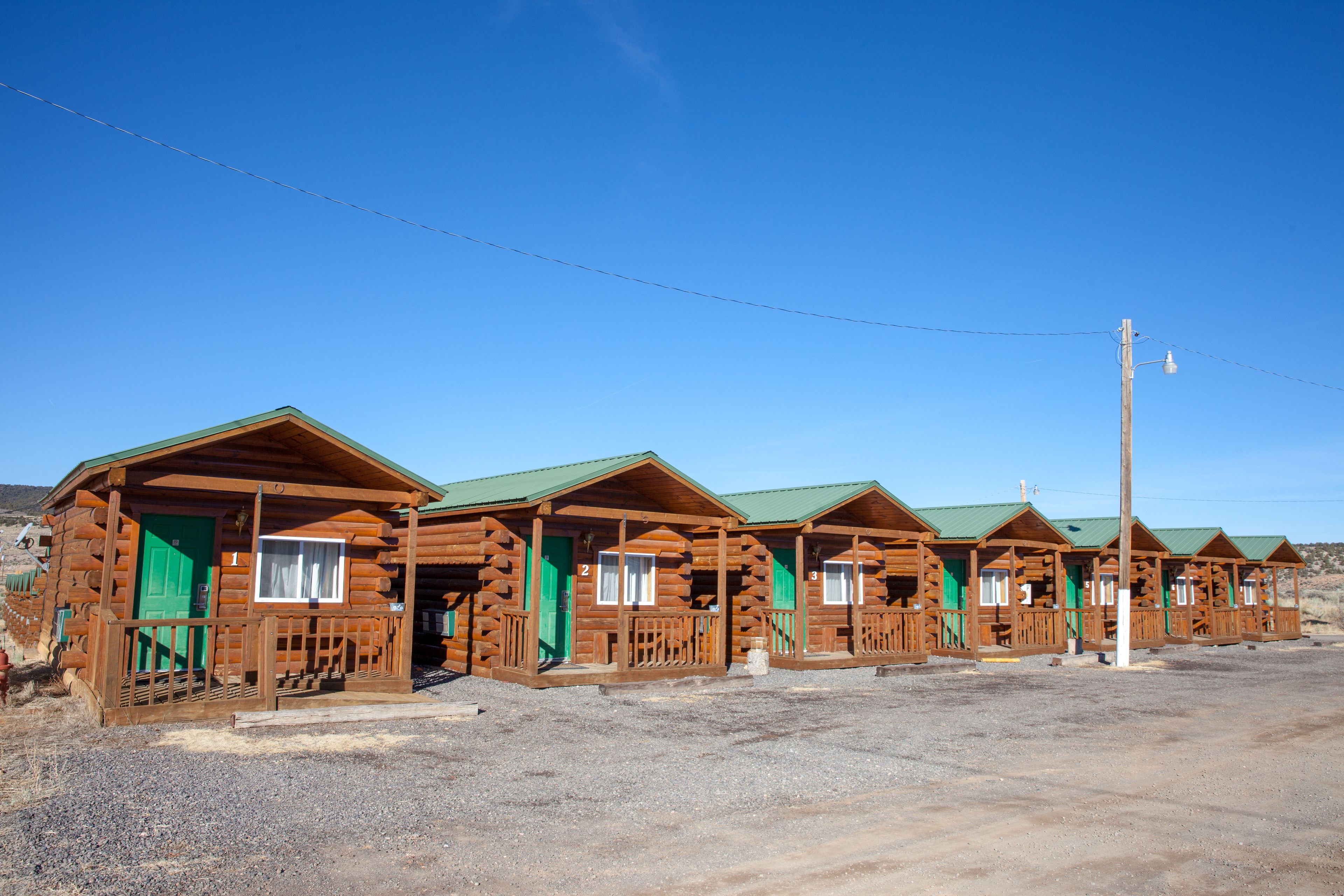 A row of log cabins with green doors and windows, arranged in a row on a gravel ground.