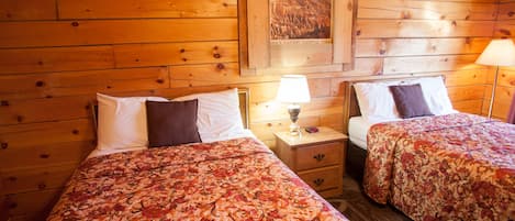 Habitación de cabaña de madera con dos camas, cada una con ropa de cama floral, una mesita de noche con lámpara y un cuadro en la pared.