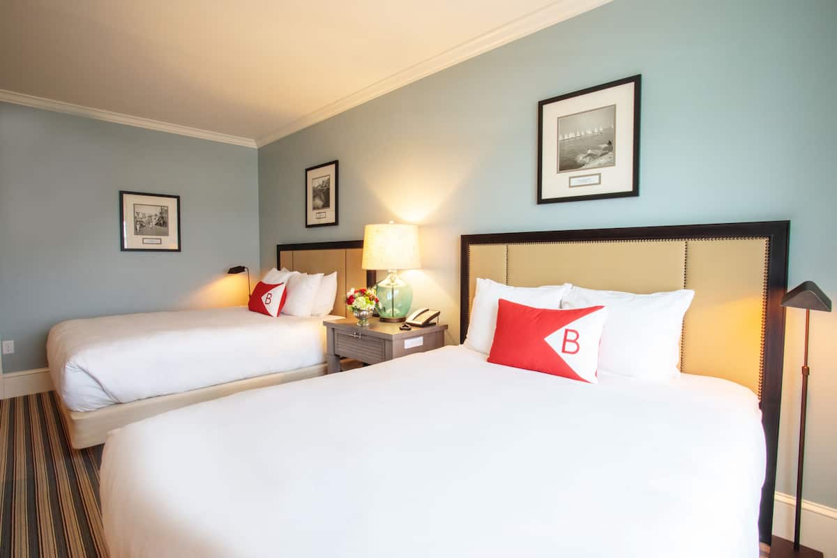 Habitación de hotel con dos camas, cada una con ropa de cama blanca y almohadas rojas con la letra 'B'. Hay una mesita de noche con una lámpara, un florero con flores y un cuadro en la pared.