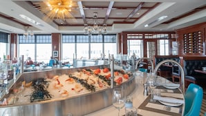 Exhibición de mariscos con camarones y algas marinas, una zona de bar con vasos y botellas, y vistas de la ciudad a través de amplios ventanales.