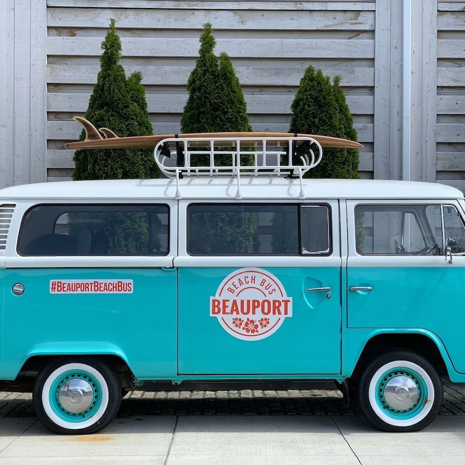 A turquoise and white vintage van with a surfboard rack, parked in front of a wooden house with potted trees.