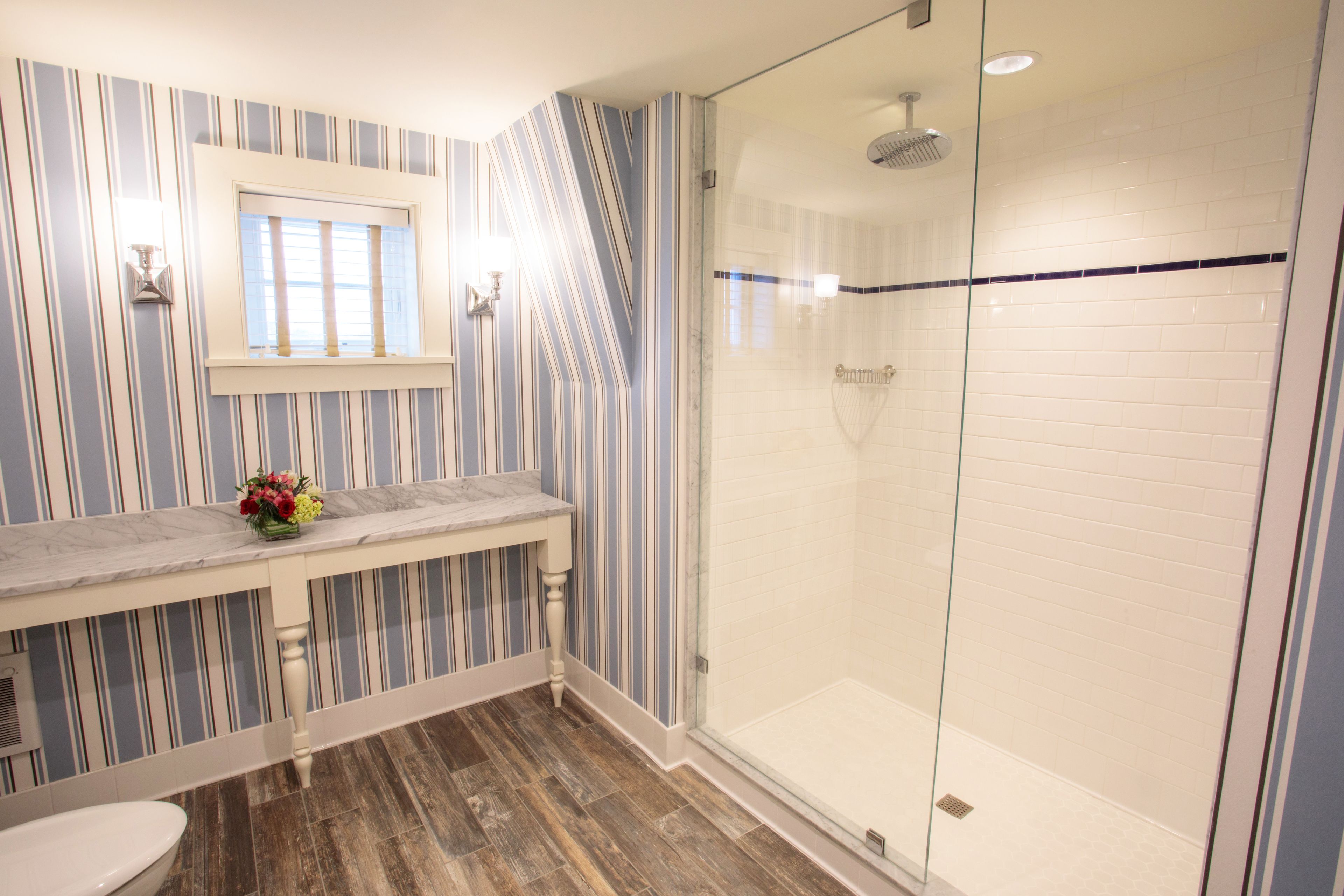 A bathroom with striped wallpaper, a marble countertop, a glass shower enclosure, and a wooden floor.