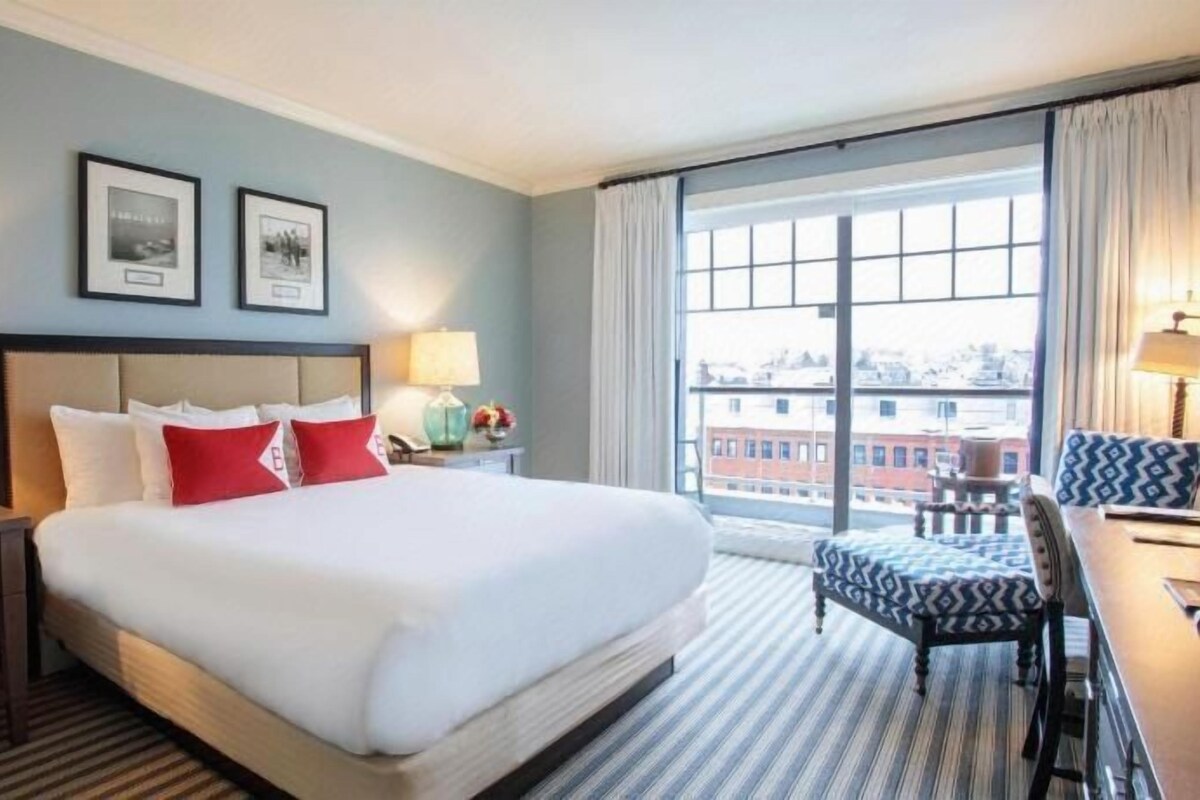 Ein Hotelzimmer mit einem großen Bett, zwei Nachttischen mit Lampen, einem Schreibtisch mit Stuhl und einem großen Fenster mit Blick auf die Stadt.