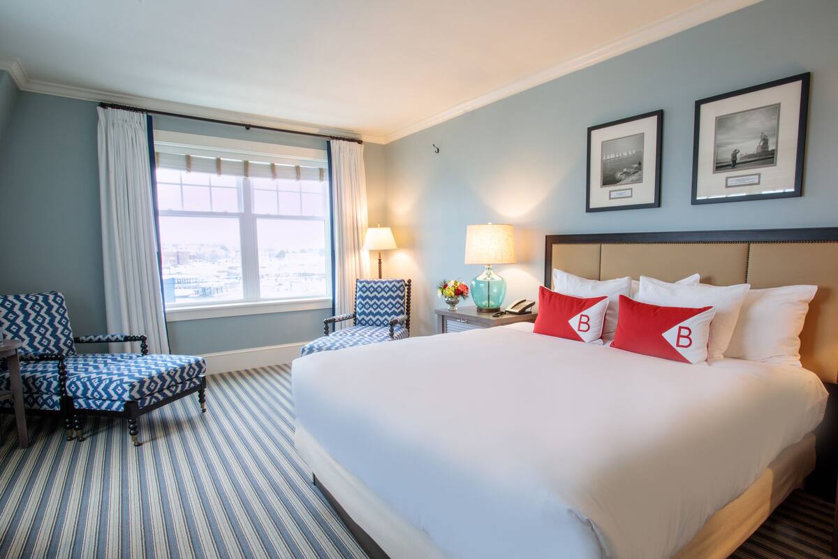 Habitación de hotel con una cama grande, dos sillas, una mesita de noche con lámpara y un ventanal con cortinas.