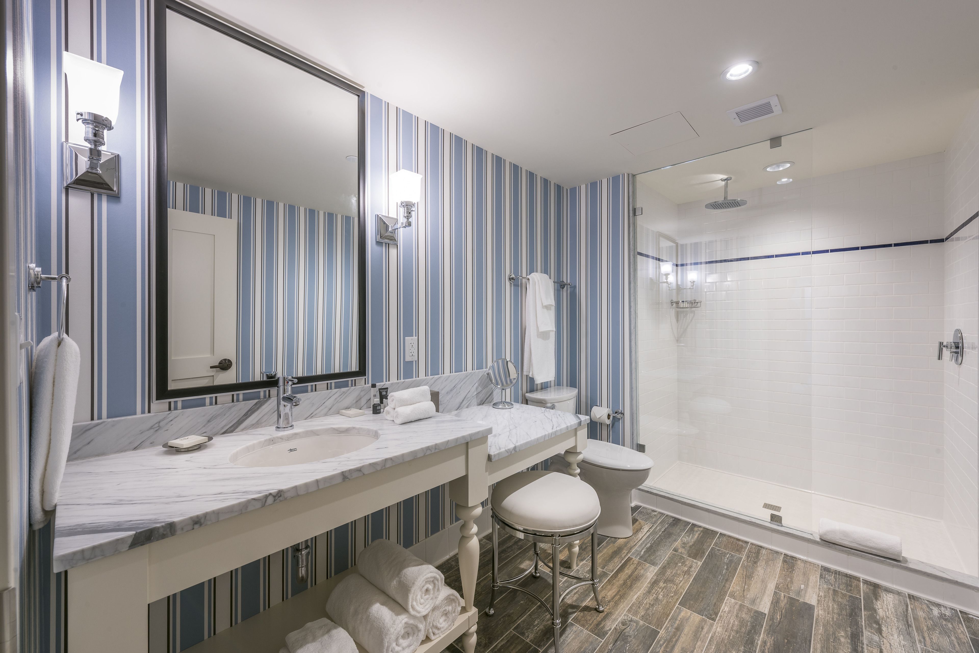 A bathroom with striped wallpaper, a marble sink, a large mirror, a toilet, and a glass-enclosed shower.