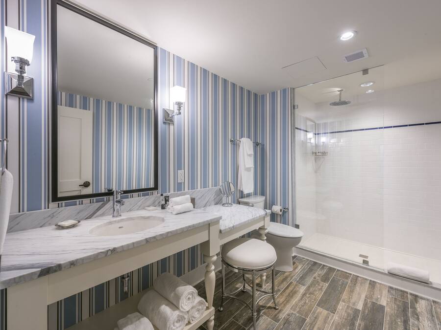A bathroom with striped wallpaper, a marble sink, a large mirror, a toilet, and a glass-enclosed shower.