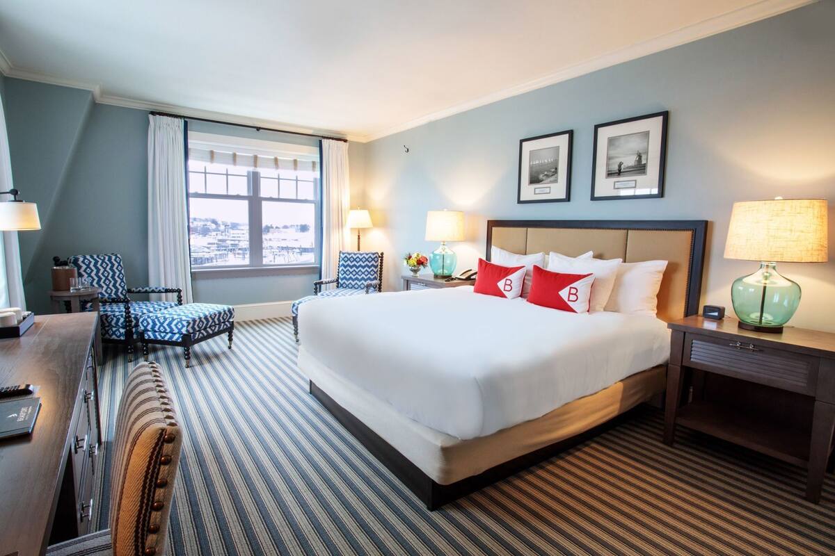 Una habitación de hotel con una cama grande, dos lámparas de noche, una silla, un escritorio y un ventanal con cortinas.