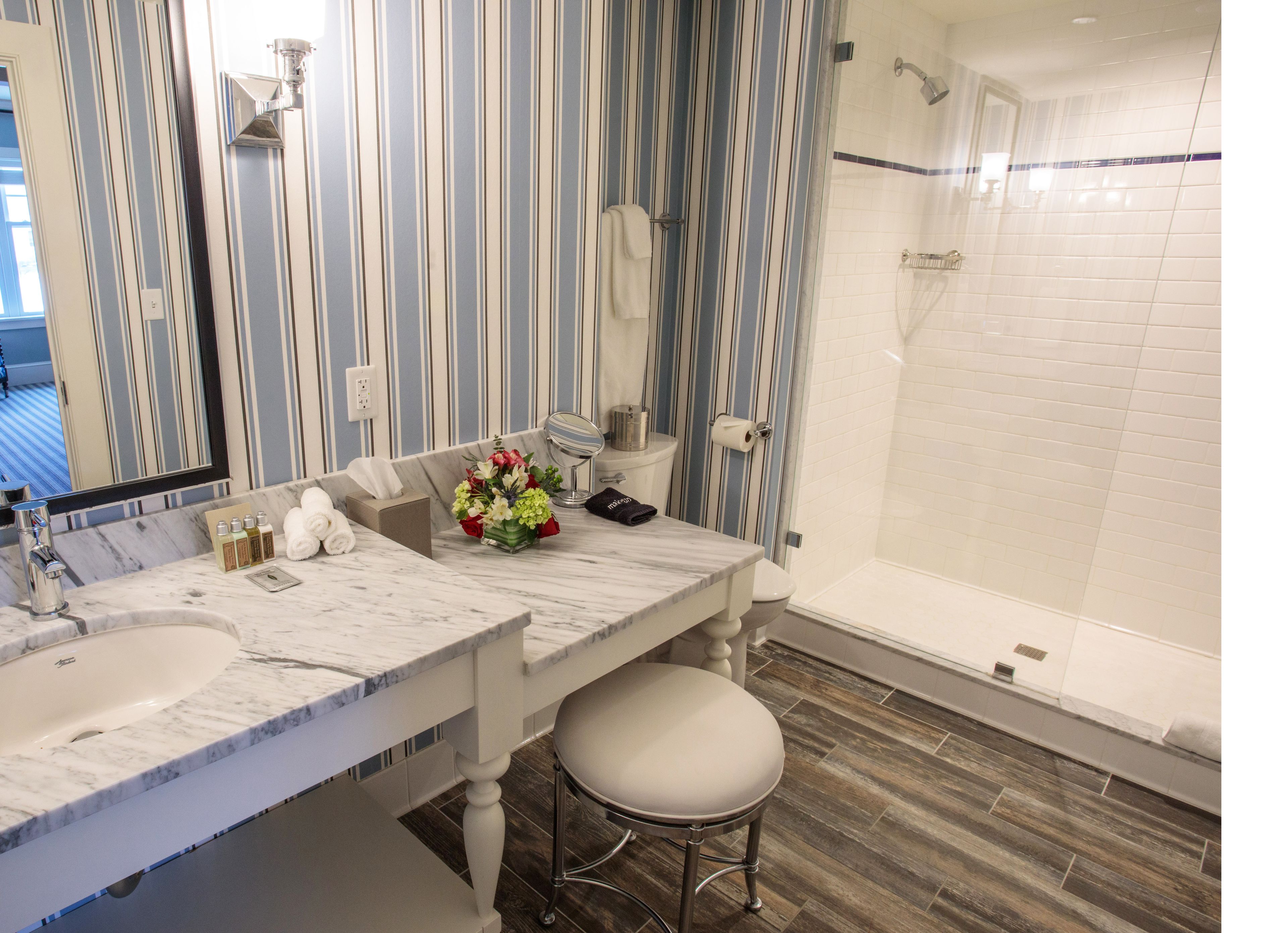 A bathroom with striped wallpaper, a marble sink, a toilet, a shower, and a stool.