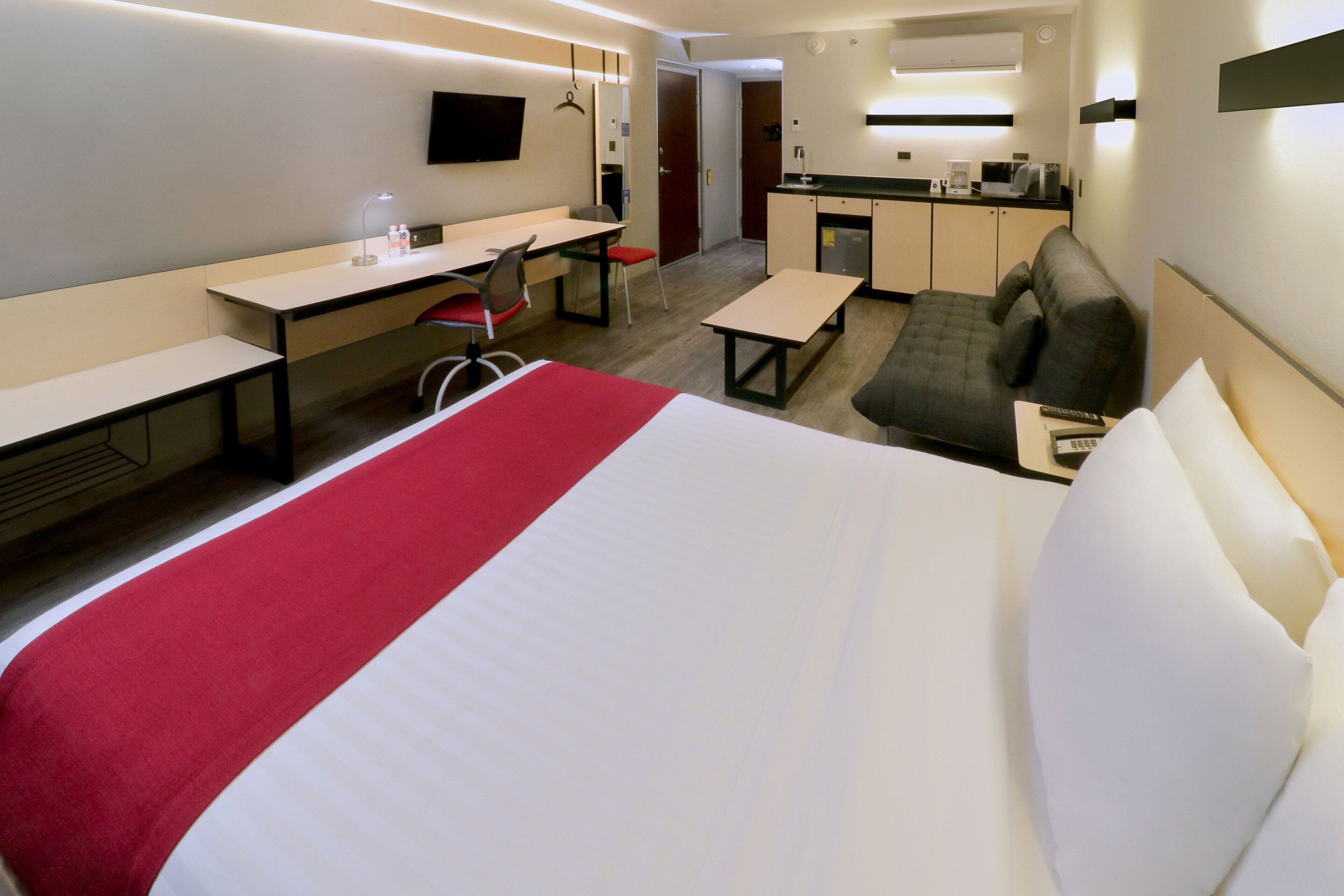 A hotel room with a bed, desk, chair, kitchenette, and a TV.