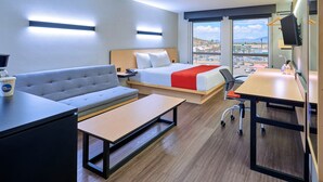 A hotel room with a bed, sofa, desk, and a view of the city. - City Express by Marriott Piedras Negras (Piedras Negras)