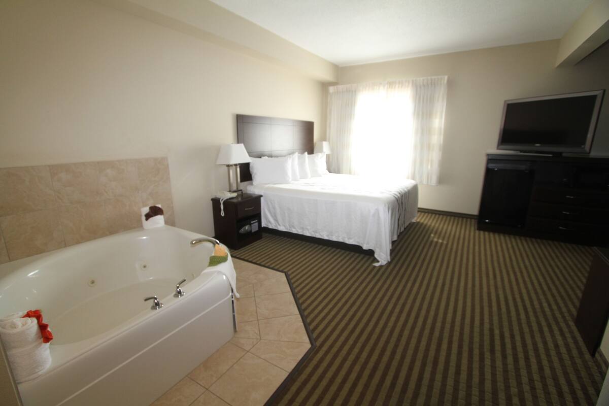 A hotel room with a large bathtub, a bed with white linens, a television, and a desk with a lamp.