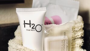 Eine Auswahl an Pflegeprodukten, darunter eine Tube mit dem Aufdruck 'H2O Therapy' und eine Seife namens 'Fresh Aloe Cleansing Bar', auf einem gemusterten weißen Handtuch.