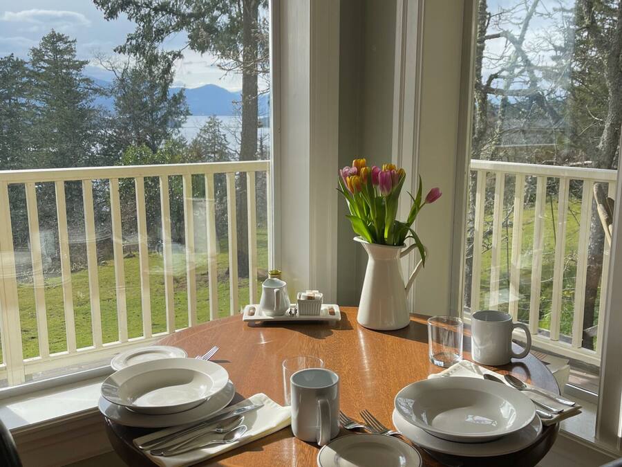 Une table à manger dressée pour quatre personnes, avec des assiettes, des couverts et un vase rempli de fleurs, sur un balcon offrant une vue sur un espace verdoyant et des arbres.