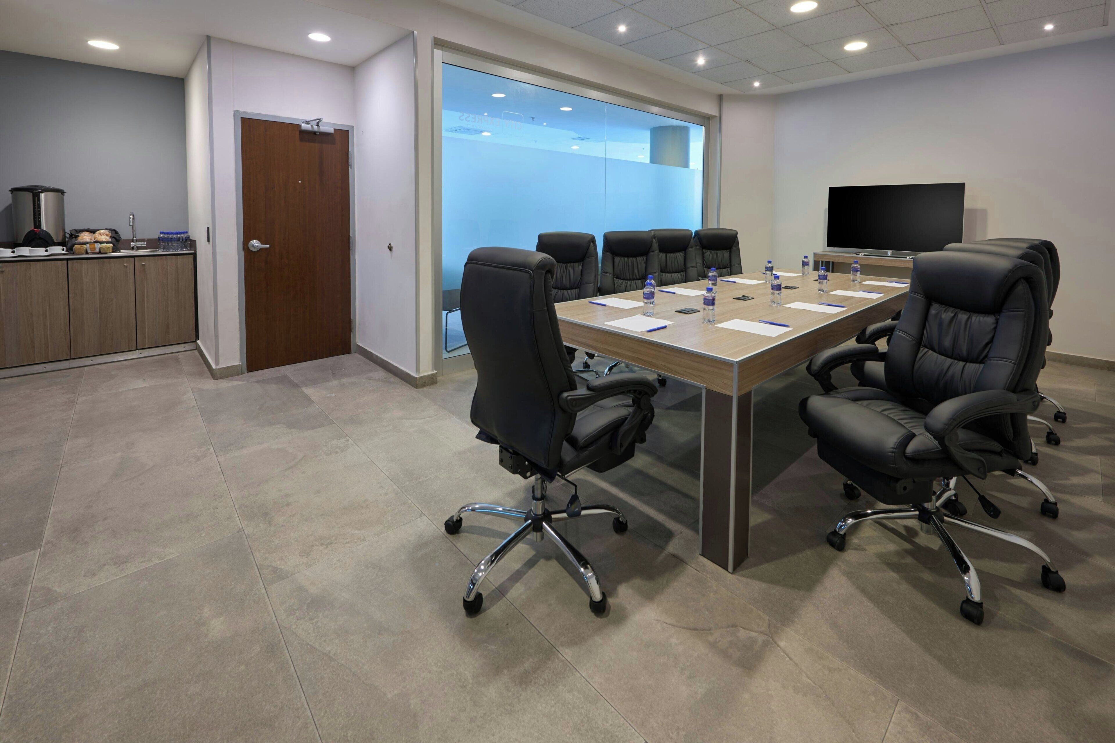 A conference room with a long table, four black office chairs, a television, and a kitchen area with a coffee maker and cabinets.