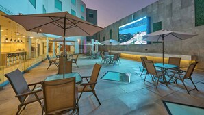 An outdoor seating area with tables and chairs, umbrellas, and a large screen displaying a scenic view. - City Express by Marriott Ciudad de México Aeropuerto (Mexico City)