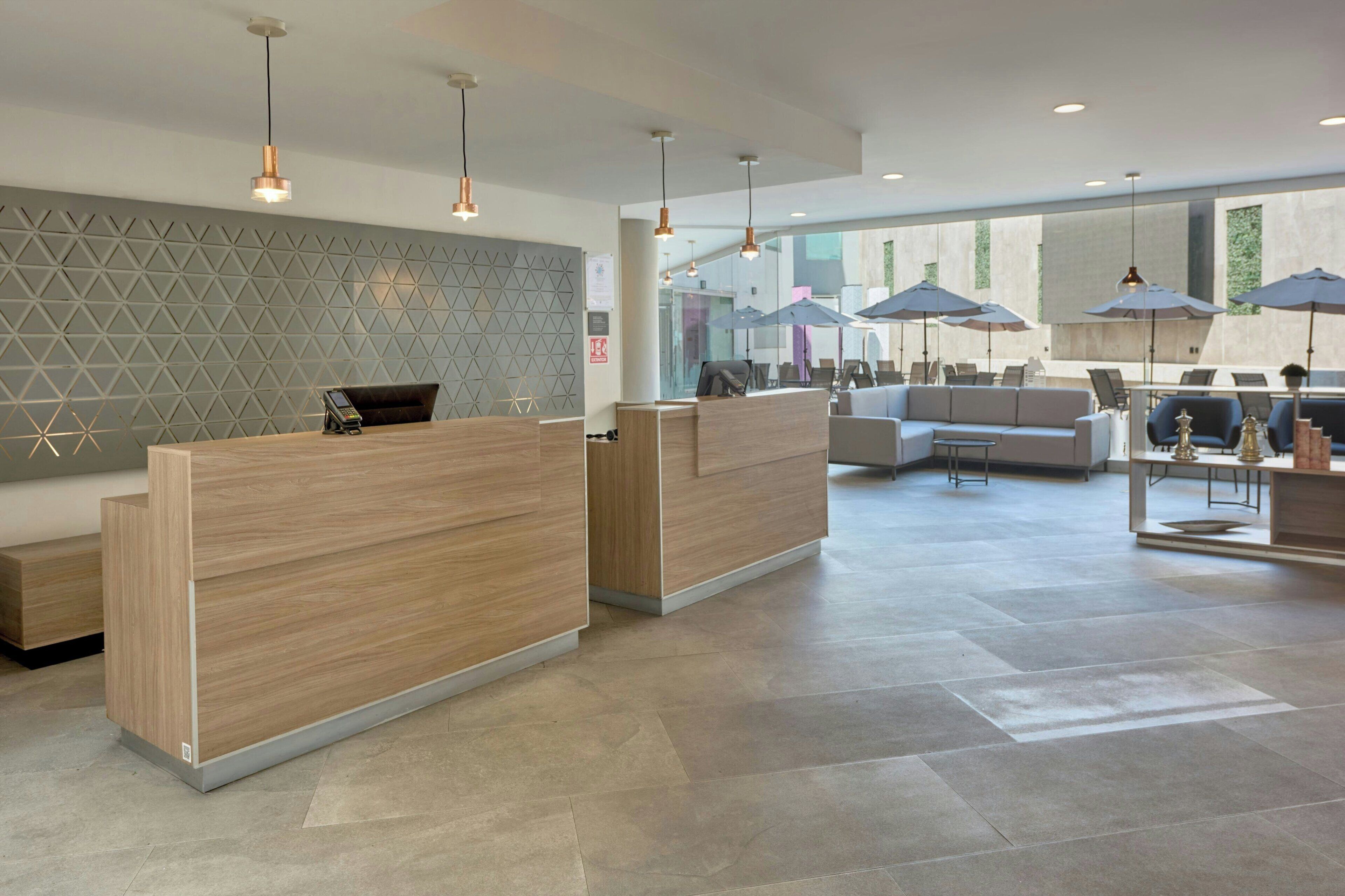 Un área de recepción moderna con un mostrador de madera, una zona de estar con sofá y ventanas amplias que dejan entrar luz natural.