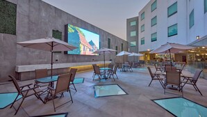 An outdoor patio area with tables and chairs, umbrellas, and a large screen displaying a scenic image. - City Express by Marriott Ciudad de México Aeropuerto (Mexico City)