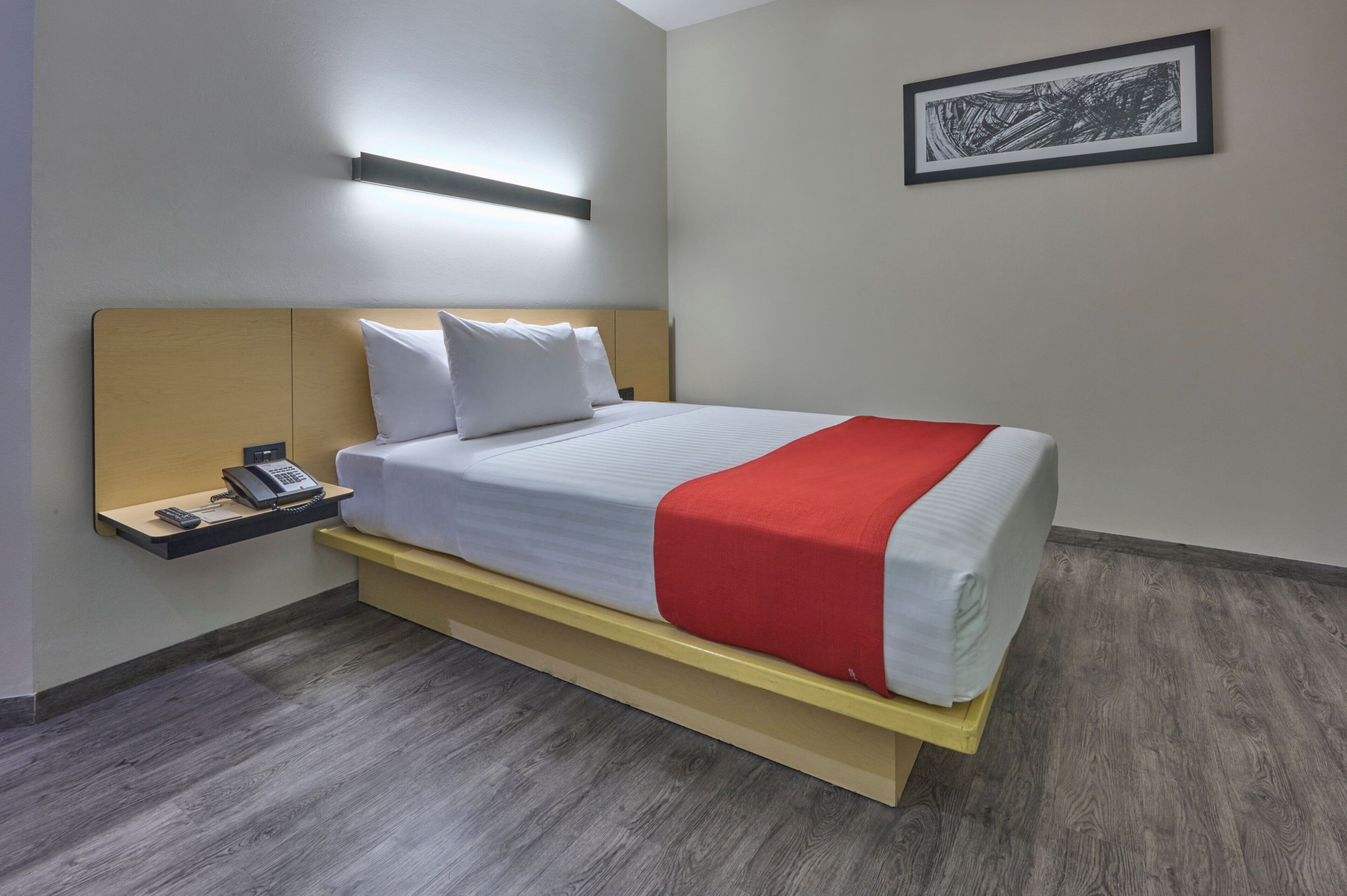 Een hotelkamer met een bed, een nachtkastje met een telefoon, een wandlamp en een ingelijst schilderij aan de muur.
