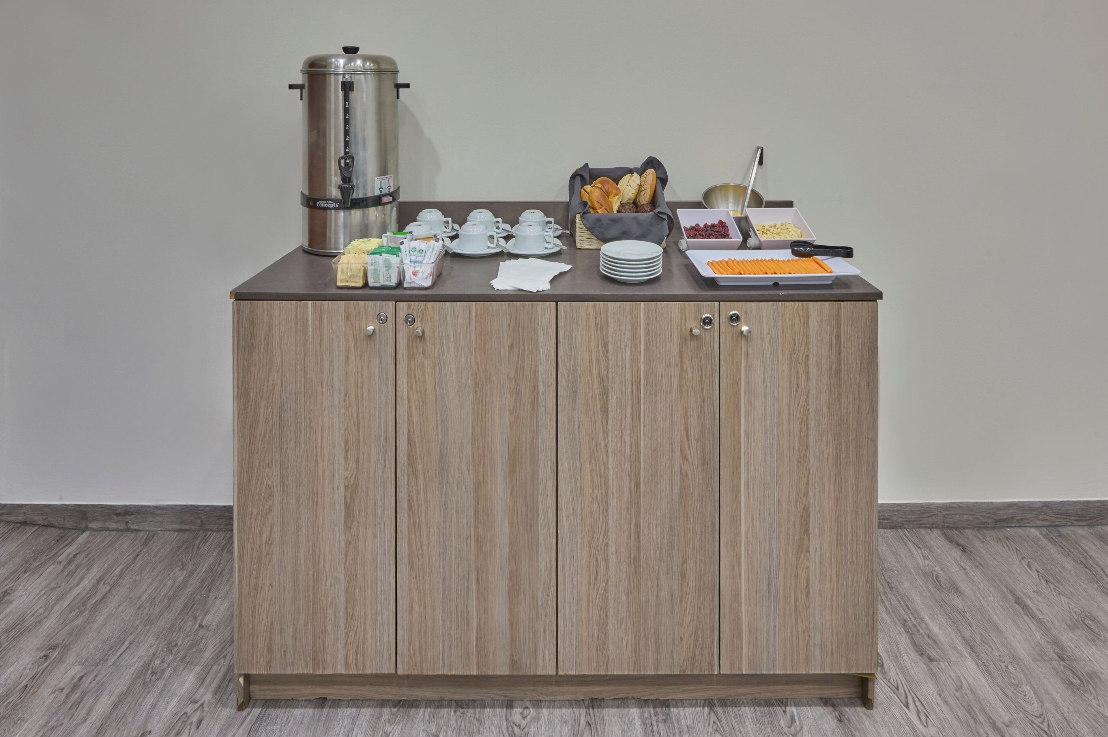 Ein Holzschrank mit Kaffeemaschine, Teekannen, Tellern, einem Brotkorb und einer Obstplatte.
