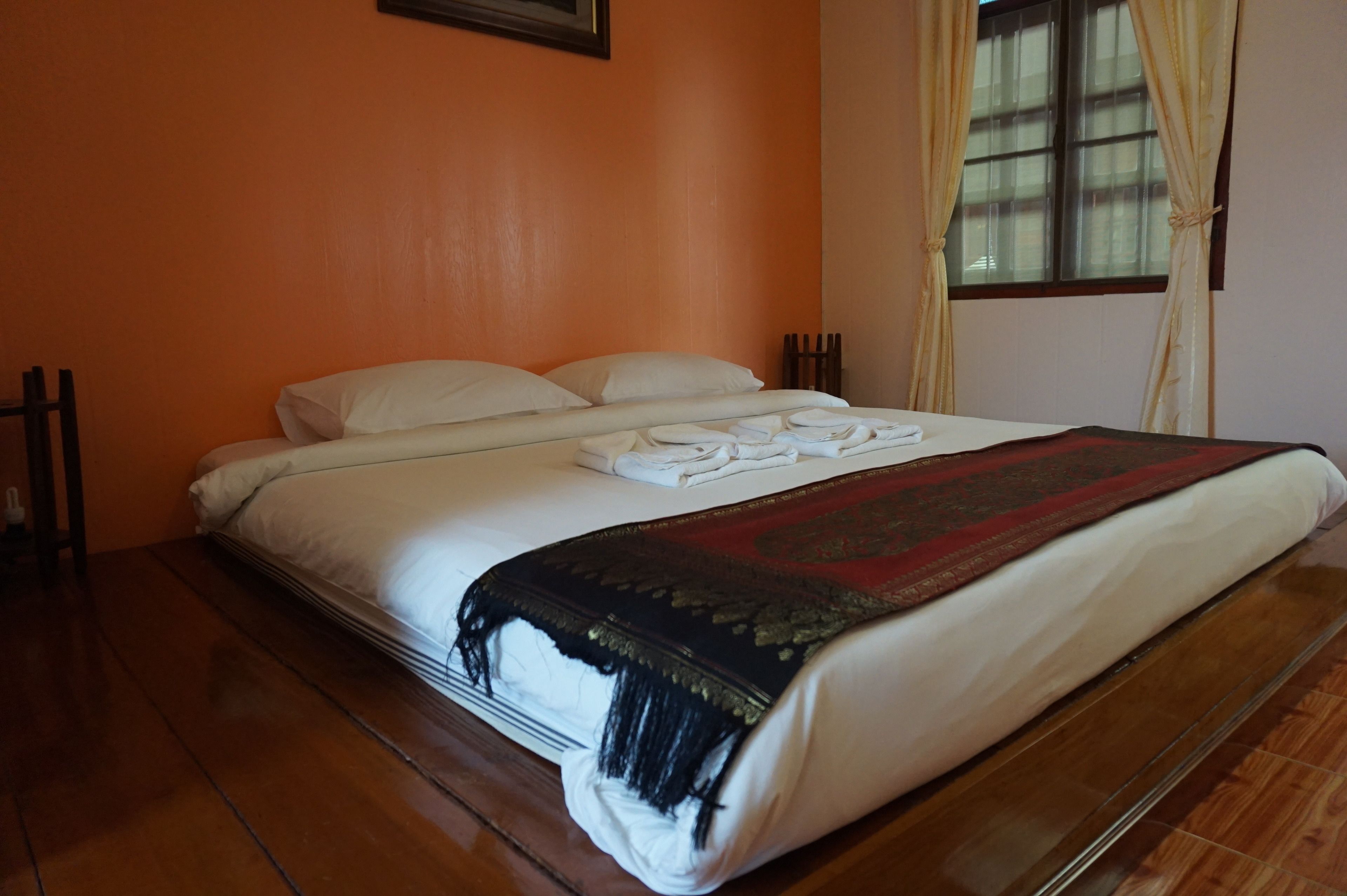 A neatly made bed with white linens and a dark patterned throw blanket, a wooden floor, and a window with curtains.
