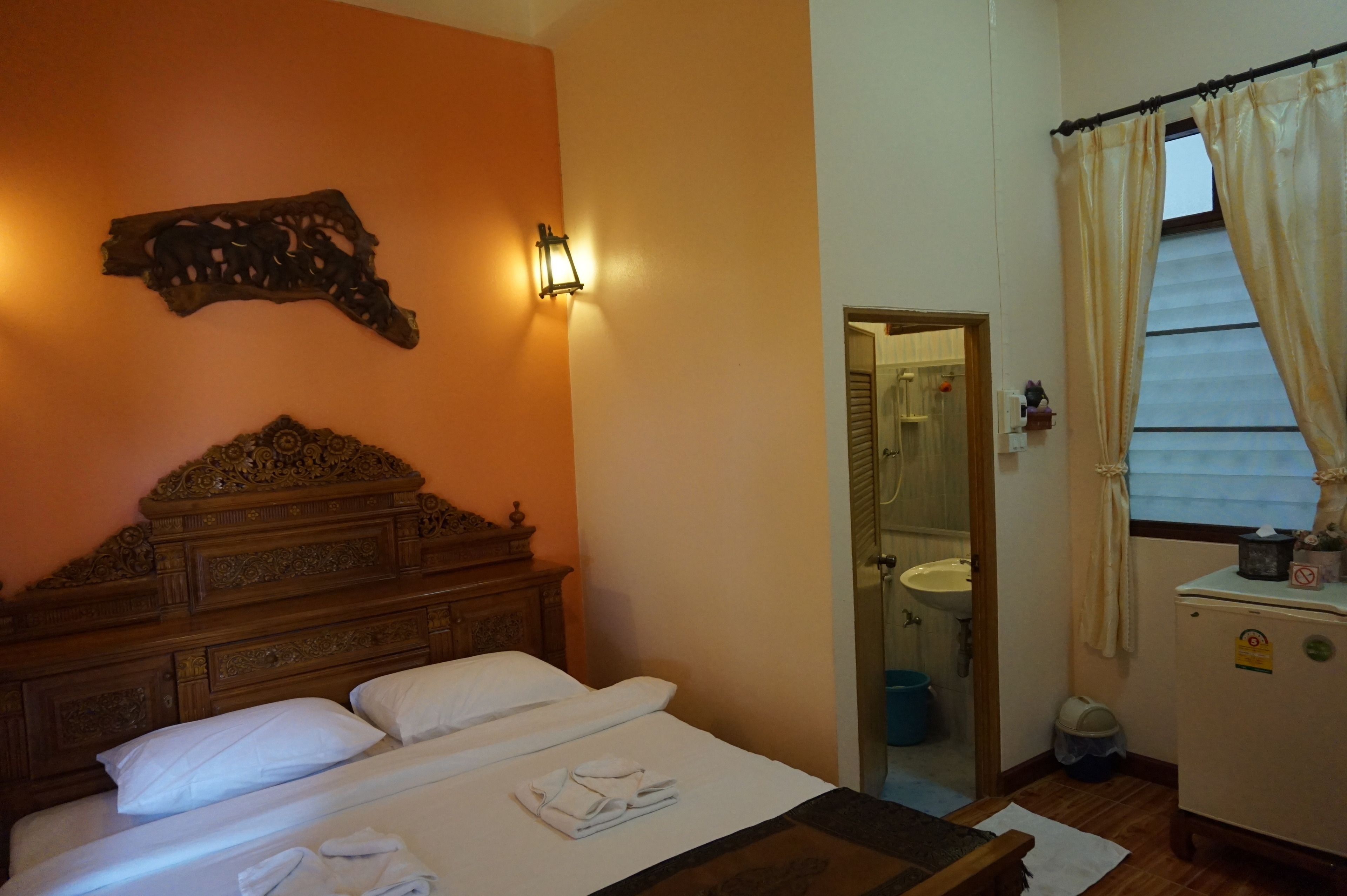 A hotel room with a wooden headboard, a bed with white linens, a wall-mounted light fixture, a mirror, and a window with curtains.