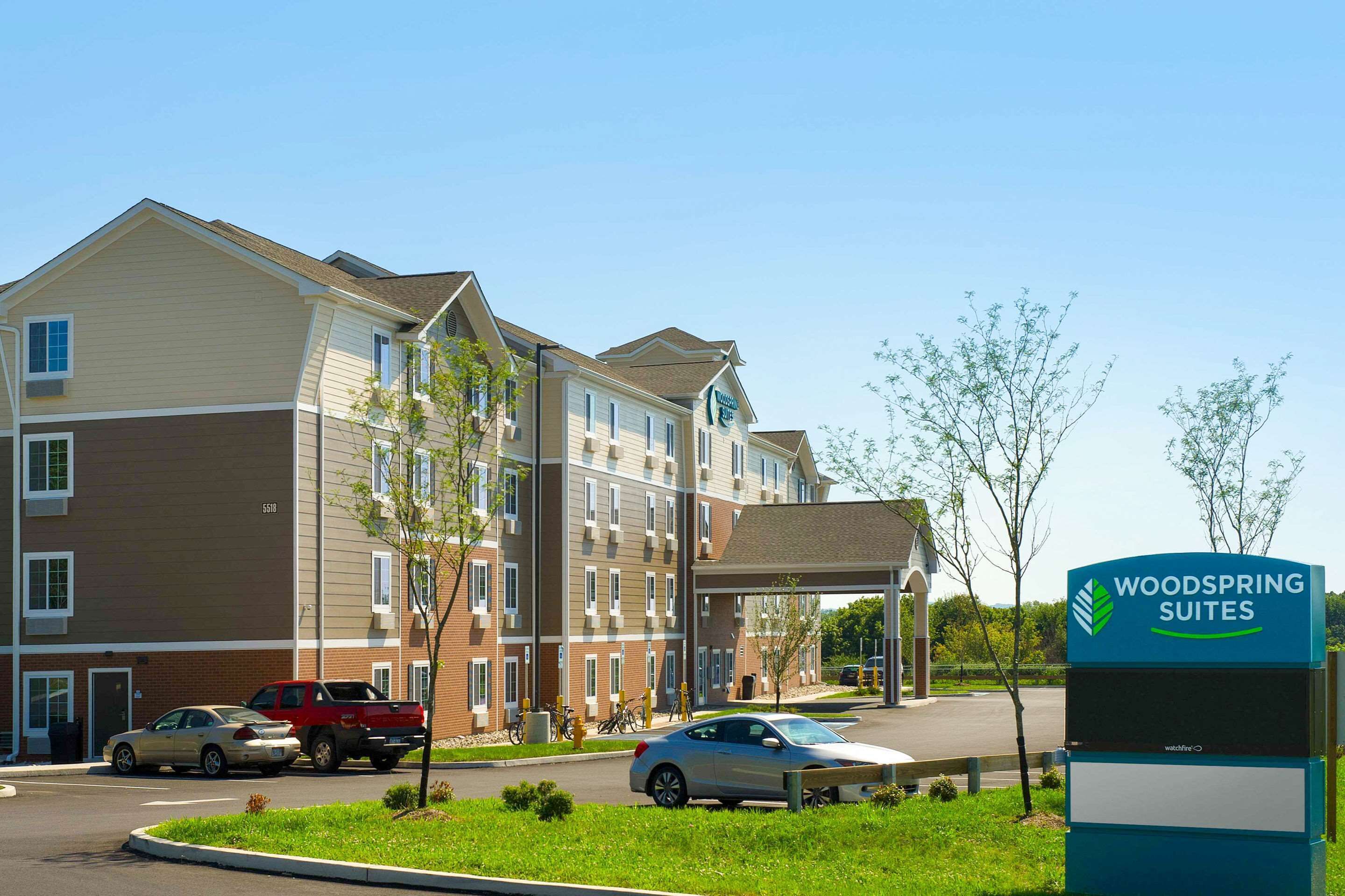 A multi-story hotel with a sign reading "Woodspring Suites." There are cars parked in front, and a covered walkway leading to the entrance.