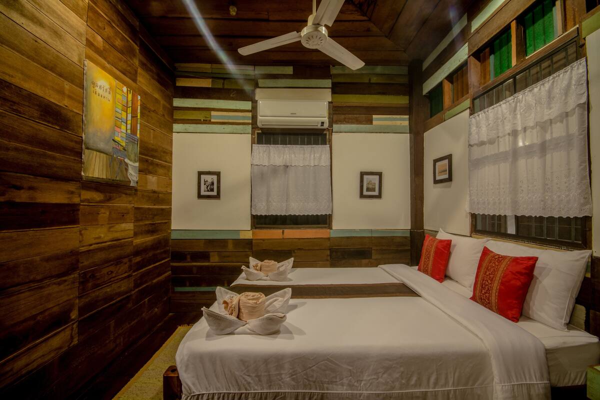 A room with wooden walls, a ceiling fan, two beds with white bedding and red pillows, and a window with lace curtains.