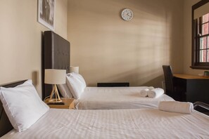 Quarto de hotel com duas camas, uma escrivaninha, uma cadeira, um relógio e um quadro na parede.