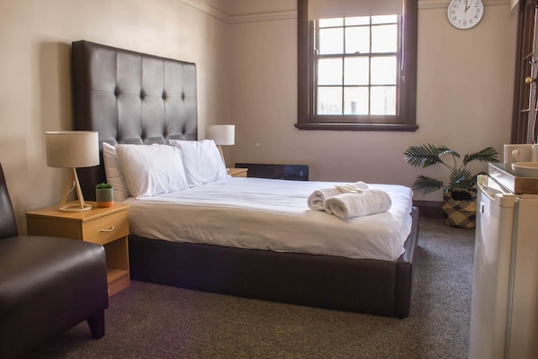 Cama bem arrumada com lençóis brancos, cabeceira escura, abajur e um pequeno vaso com planta.