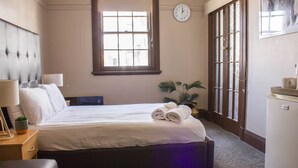 A neatly made bed with white linens and rolled towels, a bedside lamp, a small potted plant, and a clock on the wall. - Campsie Hotel (Campsie)