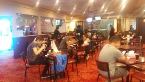 A restaurant with multiple tables and chairs, people dining, and a counter area. - Campsie Hotel (Campsie)