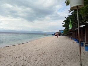 A sandy beach with a row of buildings on the right, a clear sky, and mountains in the distance. - HK Beach Resort (Moalboal)
