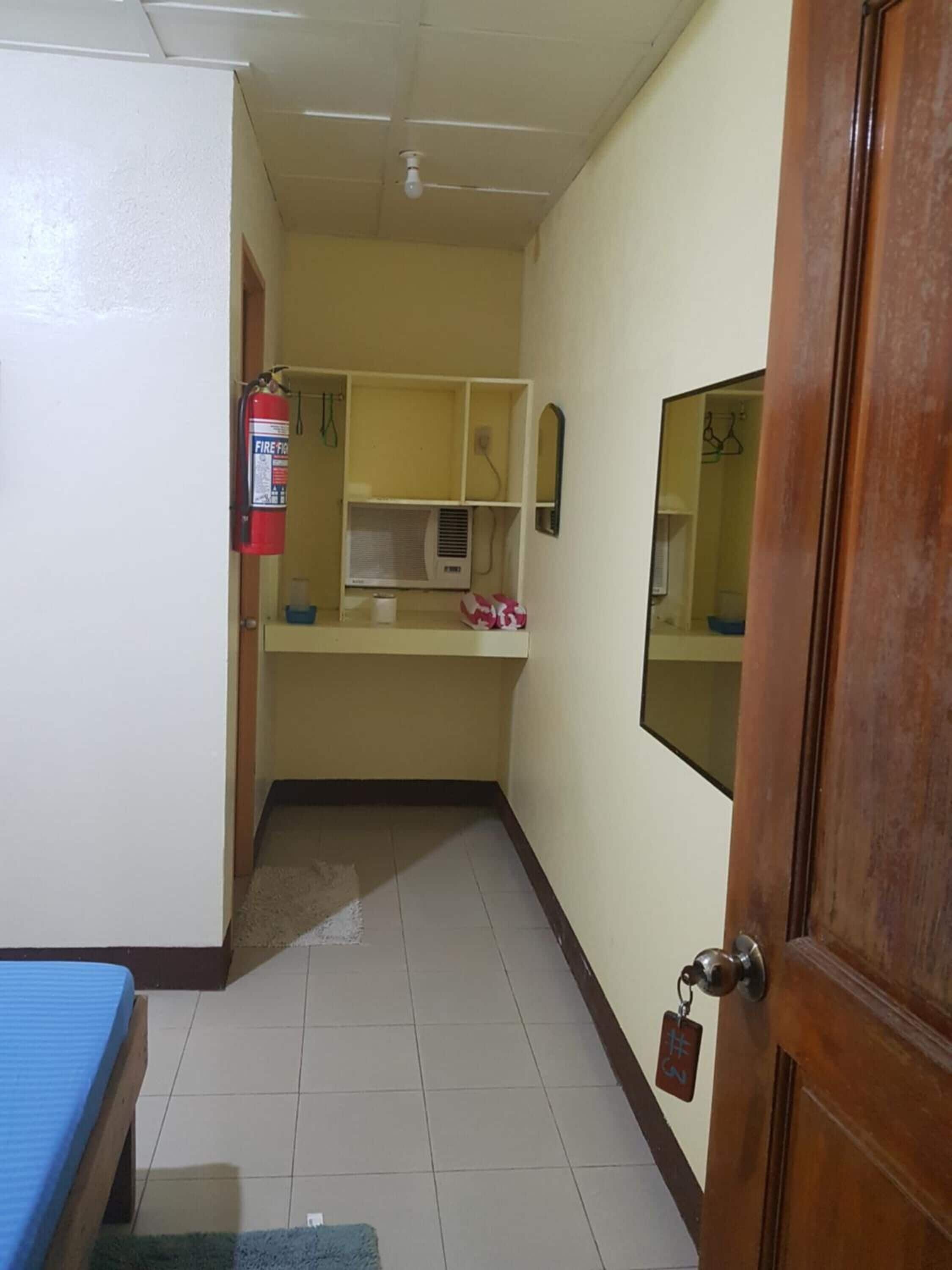 A narrow hallway with a wooden door on the right, a fire extinguisher on the left, and a small table with a blue tablecloth at the end of the hallway.