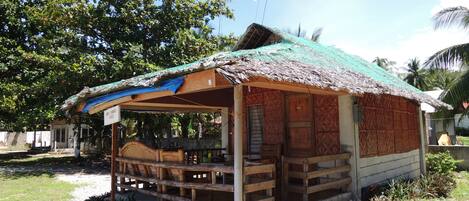 A small, rustic hut with a thatched roof and wooden walls, situated in a tropical setting with lush greenery and palm trees in the background.