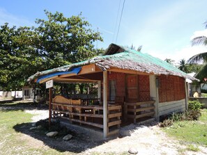 A small, rustic hut with a thatched roof and wooden walls, situated in a tropical setting with lush greenery and palm trees in the background.