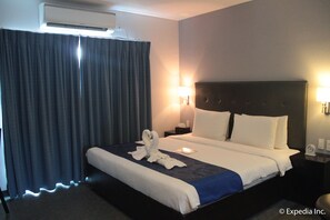 Habitación de hotel con una cama grande, cabecera, mesita de noche, lámpara y cortina.