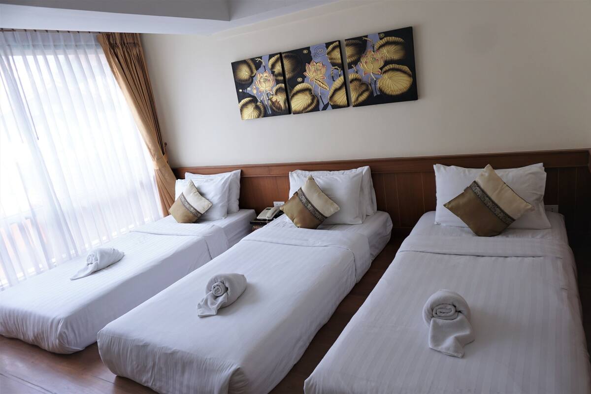 Une chambre d’hôtel avec trois lits simples, chacun muni d’un oreiller et d’une serviette roulée. Un tableau est accroché au mur au-dessus des lits, et il y a une fenêtre avec des rideaux.