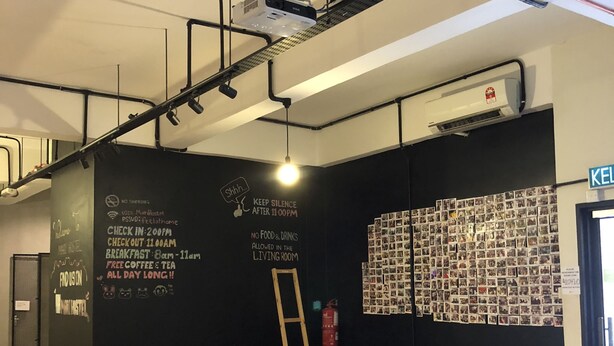 A modern lounge area with a black wall featuring various notices, a projector mounted on the ceiling, and a wall of photos. - Mari Hostel (Ipoh)