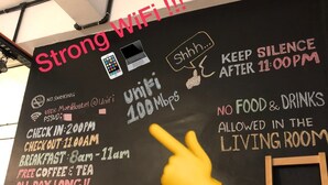 A blackboard with various rules and amenities for a hostel, including WiFi availability, check-in and check-out times, breakfast hours, and a note about no food and drinks allowed in the living room. - Mari Hostel (Ipoh)