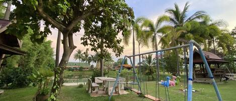 A playground with swings and a slide, surrounded by greenery and palm trees.