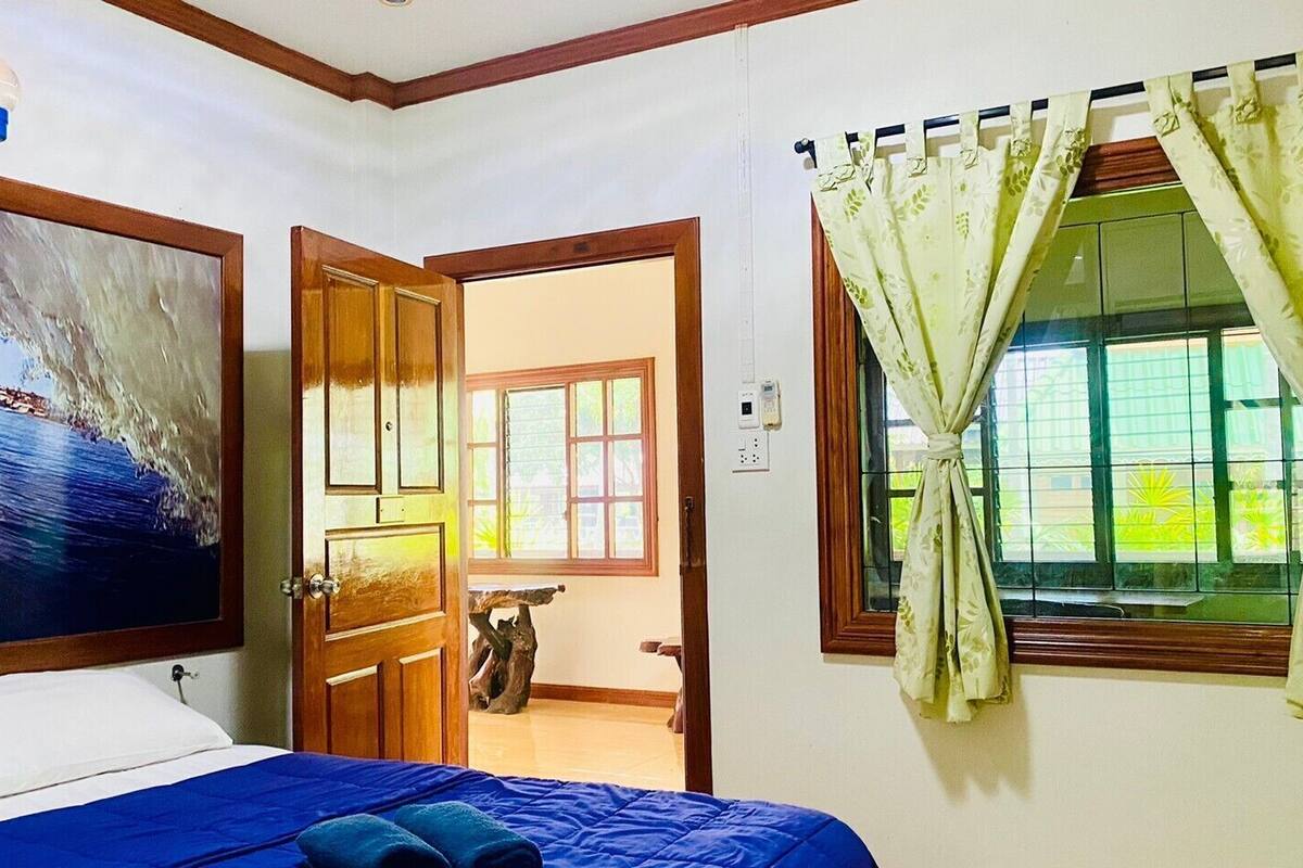 Makuuhuoneessa on sänky, taulu, puuovi, ikkuna, jossa on verhot, sekä pieni pöytä, jonka päällä on kasvi.
